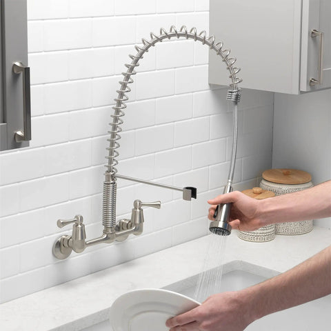 Person using Dual Handle Pro Grade Commercial Style Wall Mount Kitchen Faucet With Pull Down Nozzle And Dual Spray Options In Stainless Steel 1.8 GPM Water Saver For Heavy Duty Kitchens Perfect For Busy Kitchens And Home Chefs, rinsing dishes in a modern kitchen.