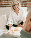 Older woman in white top smiling while presenting a wrapped gift box with a ribbon on a bed against a brick wall.