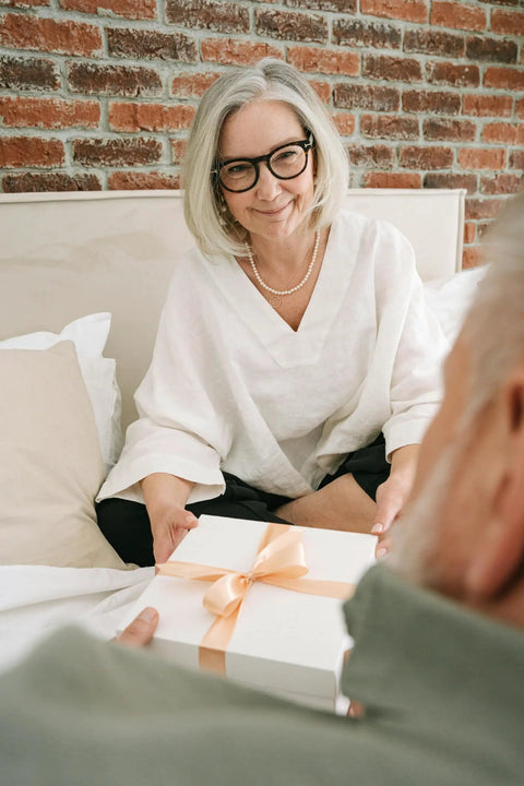 Older woman in white top smiling while presenting a wrapped gift box with a ribbon on a bed against a brick wall.