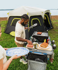 Man using Outdoor Cook Table with Adjustable Height and Foldable Four Foot Work Station for BBQ near tent by lake