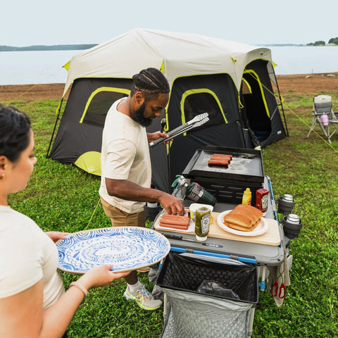 Man using Outdoor Cook Table with Adjustable Height and Foldable Four Foot Work Station for BBQ near tent by lake