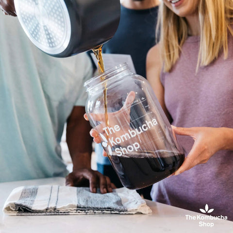 Individuals pouring tea into a glass jar for Organic 1 Gallon Kombucha Starter Kit With SCOBY Fermentation Jar Tea And Sugar Bottling Essentials And Clear At Home Brewing Instructions For Beginners To Kickstart Your Brew Like A Pro.