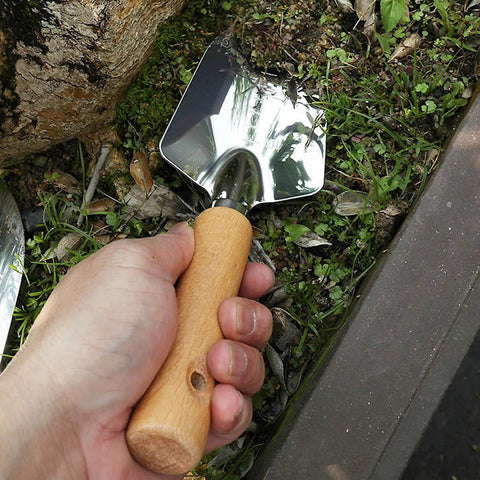Hand holding Built To Last Stainless Steel Garden Shovel With Ergonomic Wooden Handle for digging in soil near stone