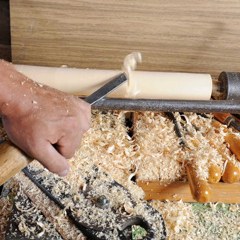 Woodturning lathe in operation with a metal chisel shaping a wooden spindle; wood shavings all around.