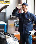 Auto mechanic in uniform standing next to an open car hood in a garage, wearing gloves and a cap, smiling at the camera.