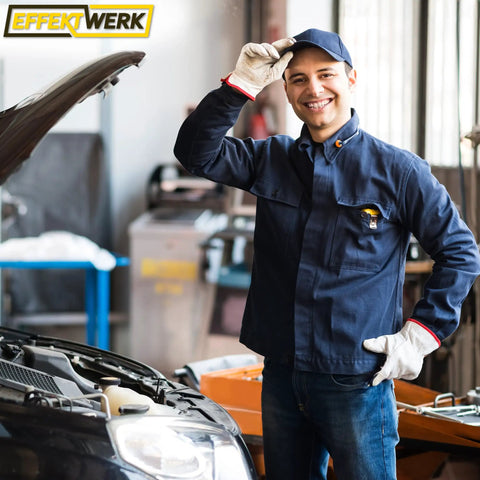 Auto mechanic in uniform standing next to an open car hood in a garage, wearing gloves and a cap, smiling at the camera.