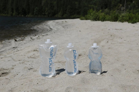 Three ultralight two-liter collapsible water bottles on sandy beach, ideal for camping and hiking, BPA free, rolls up when empty.