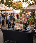 Outdoor market stall with a woman displaying jewelry on a black tablecloth, under string lights, with plants and shoppers in background.