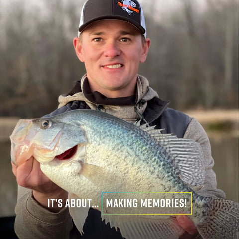 Person holding a large crappie fish by a lake, wearing a cap and jacket, emphasizing fishing memories.