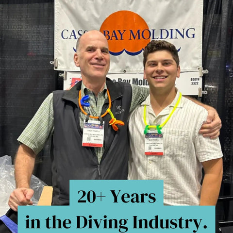 Two people smiling and posing at a diving industry booth, wearing colorful dive mouthpieces around their necks.