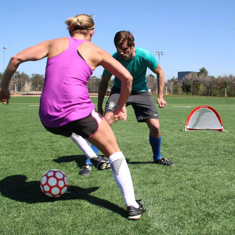 Two people playing soccer on grass with Portable Pop Up Soccer Goals For Backyard Set Of Two 4 Foot Nets visible in background