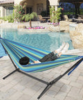 Person relaxing in an extra long and wide double hammock with blue green stripes by a pool, cotton weave, 94 by 63 inch bed.