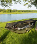 Person resting in Bug Free Camping Hammock With Mosquito Net, Ultralight Portable Outdoor Hammock, set up between trees near water.