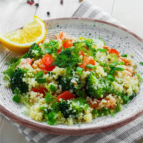 Bowl of couscous salad with broccoli, tomatoes, and dill, garnished with a lemon wedge on a checkered cloth napkin.