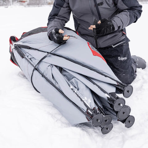 User preparing the Quickfish Six Person Portable Pop Up Ice Fishing Shelter With Fully Quilted Insulation No Trip Door And Deluxe Anchors For Hell Of A Winter Setup In Red Black on snow.