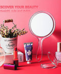 Double Sided Handheld Mirror With 1X And 2X Magnification Clear Handle Round Makeup Mirror For Face Detailing And Mascara Application Great For On The Go, shown with handle facing down on pink background among beauty products.