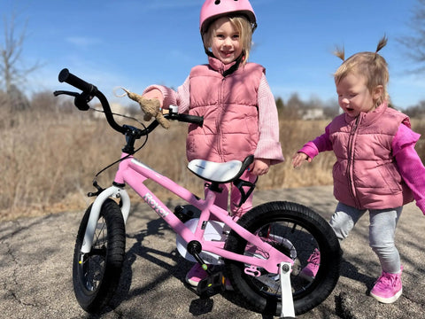 Two young girls with a 12 Inch Kids Bike For Toddlers Girls Learning Bicycle With Training Wheels Easy Peasy Assembly Safe Handbrake Front Caliper Brakes Ideal Beginner Ride For Ages 3 To 4 Pink on a sunny day.