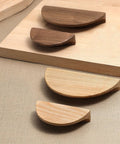 Variety of semi-circular wooden cabinet drawer pulls displayed on a textured surface, showing walnut and lighter wood finishes.