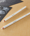 Two white charcoal pencils for sketching and highlights on a wooden surface, next to a black paper with white designs.