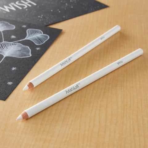 Two white charcoal pencils for sketching and highlights on a wooden surface, next to a black paper with white designs.