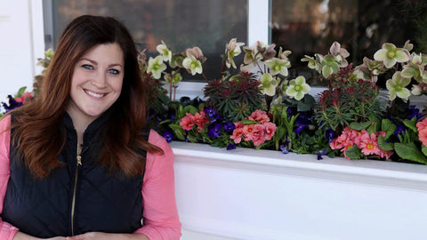 Person standing next to a window box with colorful flowers including pink and white blossoms.
