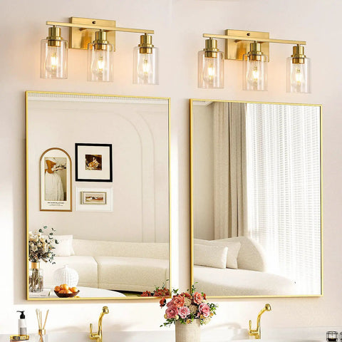 Three-Light Vanity Light With Clear Glass Shades In Brushed Brass Finish Wall Sconce Installed Above Bathroom Mirror