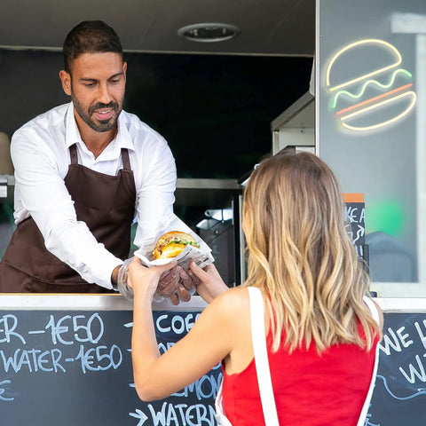 Man serving food to woman with Burger Neon Sign For Wall Decor 12x12 Inch LED Neon Light in background, cafe setting