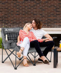 Couple sitting on Two Person Padded Camping Chair For Outdoor Lounging, Gray – Heavy Duty Oversized Camping Loveseat With Cup Holders