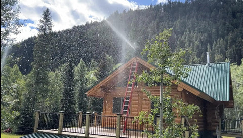 Log cabin with activated Wildfire Protection Sprinkler System With Bracket And Hardware, spraying water, forest backdrop.