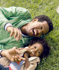 Father and child laughing on grass, showcasing Kids And Baby SPF 50 Sunscreen Spray And Bug Spray Combo, Oxybenzone And Octinoxate Free.
