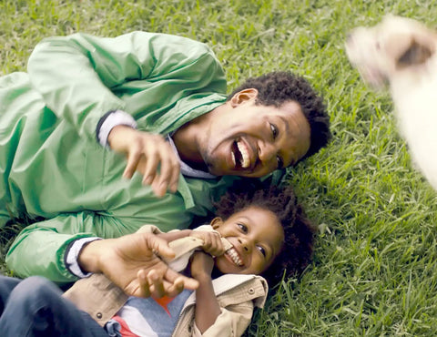 Father and child laughing on grass, showcasing Kids And Baby SPF 50 Sunscreen Spray And Bug Spray Combo, Oxybenzone And Octinoxate Free.