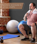 Man sitting on a chair with a towel, next to exercise balls and weights, in a gym setting.