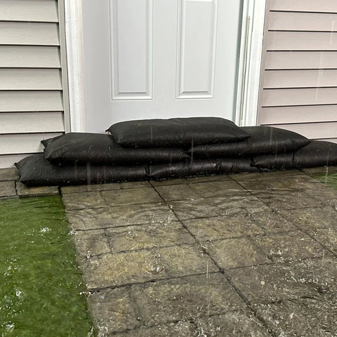 Stacked black flood bags at door showing Water Activated Flood Bags Two Pack No Sand No Labor Needed Ready To Use Expand Up To Three Point Five Inches In Minutes Protect Doorways Garages And More Lightweight And Compact For Easy Storage in use during rain