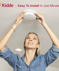 Woman installing Dual Hazard Smoke And Carbon Monoxide Detector With Voice Warning Hardwired Setup And Ten Year Sealed Lithium Battery Backup Interconnectable Network Built Tough For Zero Drama Family Safety White on ceiling.