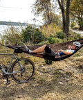 Cyclist in ultralight pocket hammock for solo adventures, tear-resistant HMPE ripstop fabric, tied between trees by a lake.