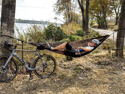 Cyclist in ultralight pocket hammock for solo adventures, tear-resistant HMPE ripstop fabric, tied between trees by a lake.