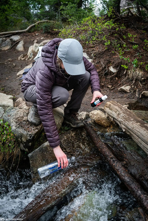 Person using Compact Palm-Sized Water Filtration System With Dual Threaded Mini Plus And Faucet Adapter For Boil Alerts White 0.1 Micron Filter Removes 99.99999% Bacteria And 99.9999% Protozoa And Cysts Plus 100% Microplastics Removal For Home Or Outdoor Faucets near a stream