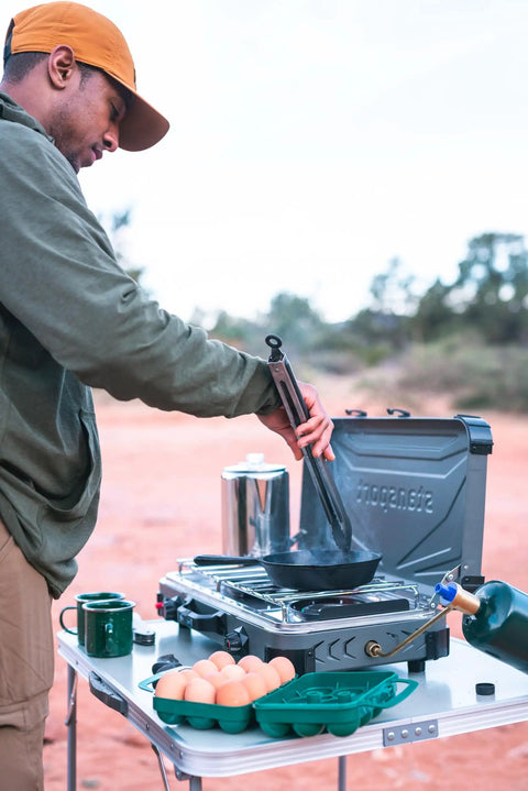 Person cooking outdoors with a Tough 12-Egg Plastic Carrier With Locking Lid, Molded Handle, And Secure Fit For Camping visible on table.