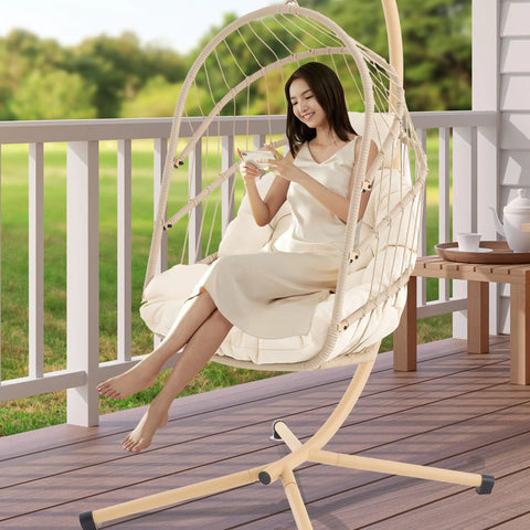 Woman sitting in Swing Egg Chair With Stand For Indoor Outdoor Use, durable wicker rattan basket with UV resistant cushions, beige