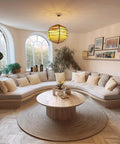 Cozy living room with a light grey modular couch, a round wooden coffee table on a woven rug, and decorative plants by arched windows.
