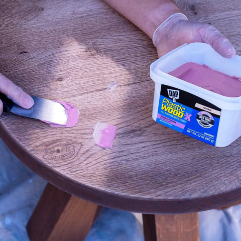Hand applying Plastic Wood Filler With DryDex Dry Time Indicator, Natural Color, 32 Oz to wooden table surface.