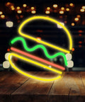 Neon burger sign glowing in yellow, green, and red against a blurred night backdrop, mounted on a wooden surface.