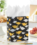 Medium Graduation Gift Bag With Tissue Paper Featuring Gold And Silver Mortarboards On Black for College High School Eighth Grade Kindergarten And Nurses Gift Ready, displayed on a kitchen counter with snacks.