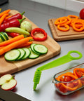 Green handheld spiral slicer, plastic kitchen tool with cucumber slices and carrot sticks on a cutting board.