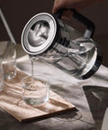 Glass Water Filter Pitcher With Stainless Steel Refillable Filter System pouring water into a glass on a kitchen table