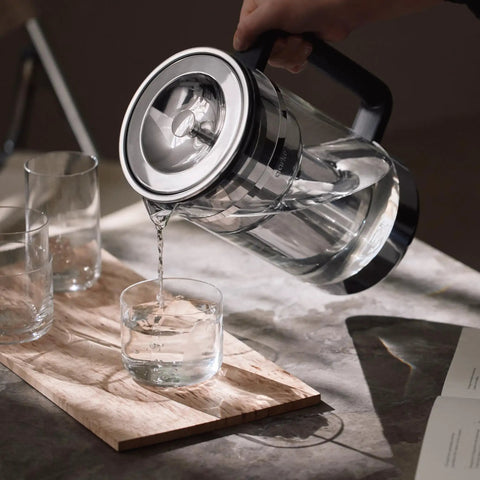 Glass Water Filter Pitcher With Stainless Steel Refillable Filter System pouring water into a glass on a kitchen table