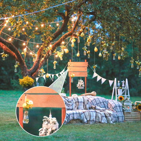 Outdoor garden scene with tree decorated with lights and hanging jars, cozy seating with plaid blanket, wooden crates, and flower arrangements.