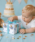 Baby reaching for a gift wrapped in Blue Teddy Bear Hot Air Balloon Baby Wrapping Paper Roll For Baby Boy Birthday And Baby Shower Mini Roll 17 Inches By 32 Point 8 Feet 46 Sq Ft Totally Cute Gift Wrap.