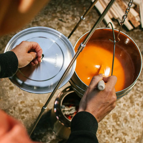 Person cooking with Campfire 2 Pot Wood Burning Camping Stove Set For 4 Plus People, showing soup in metal pot over fire.