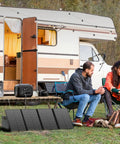 Portable Solar Generator 300W Peak Power Station With 260Wh Battery And 40W Solar Panel Included For Home Use Camping Outdoor Adventures On The Go Off Grid Backup Power in use beside a camper with people sitting around a campfire, highlighting its outdoor capability.